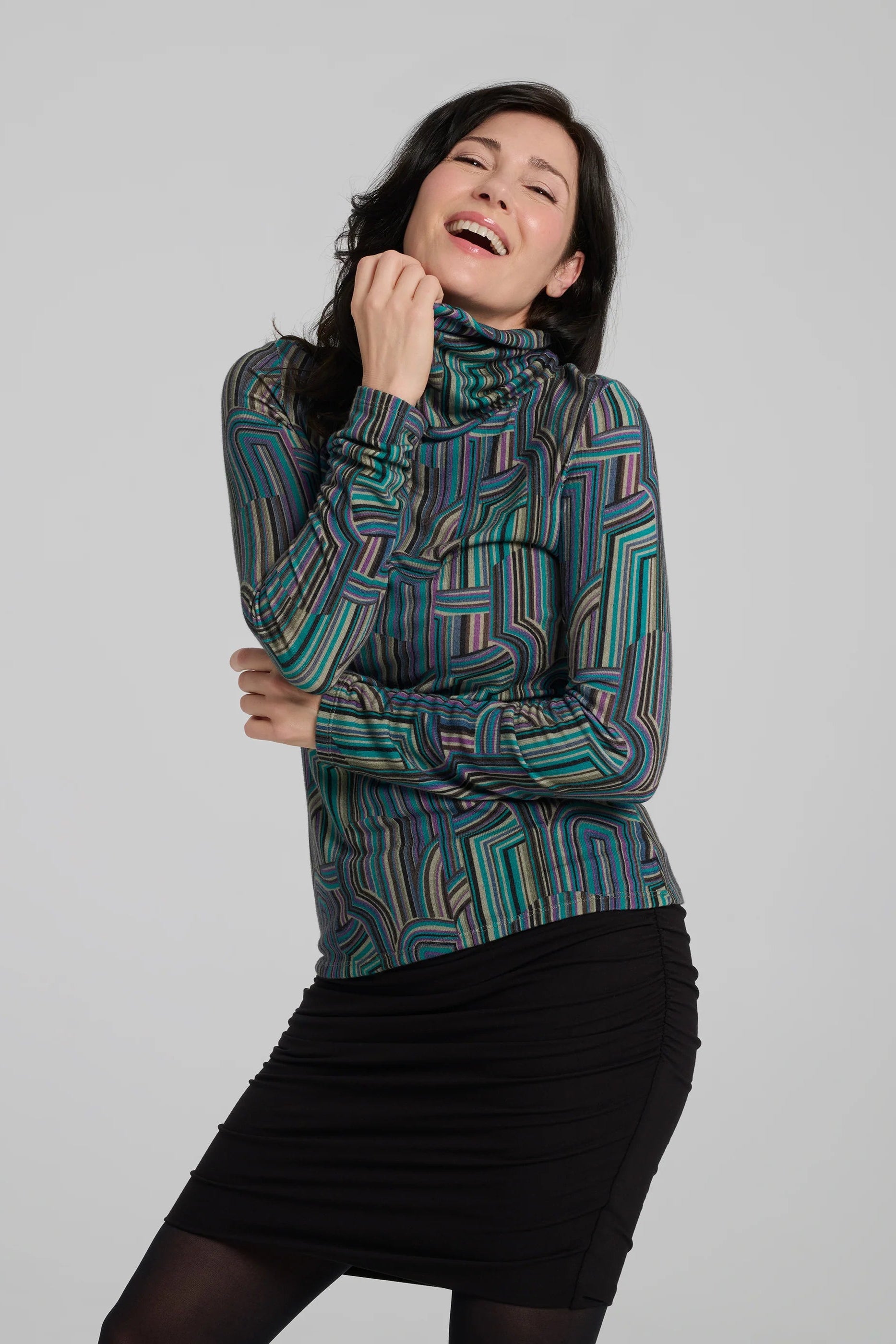 A woman wearing the Yrene Top by Kollontai in Turquoise, a geometric print turtleneck. She is wearing it with a black skirt and standing in front of a grey background. 