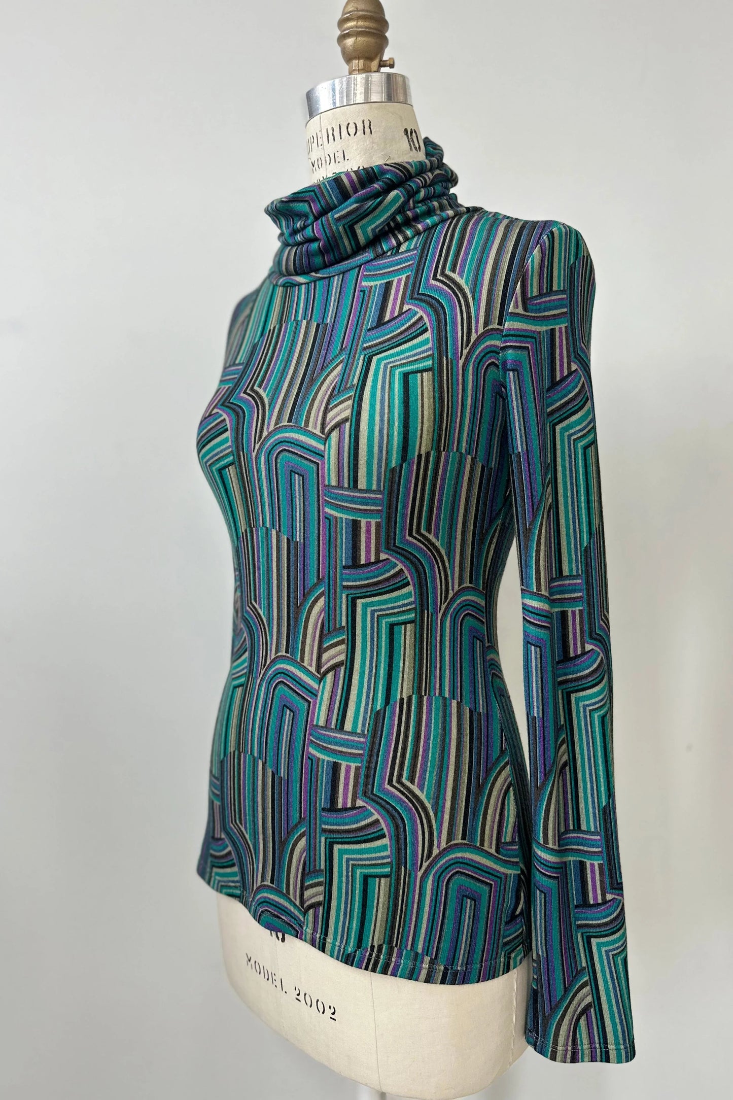 Side view of the Yrene Top by Kollontai in Turquoise, a geometric print turtleneck, is shown on a mannequin against a grey background. 