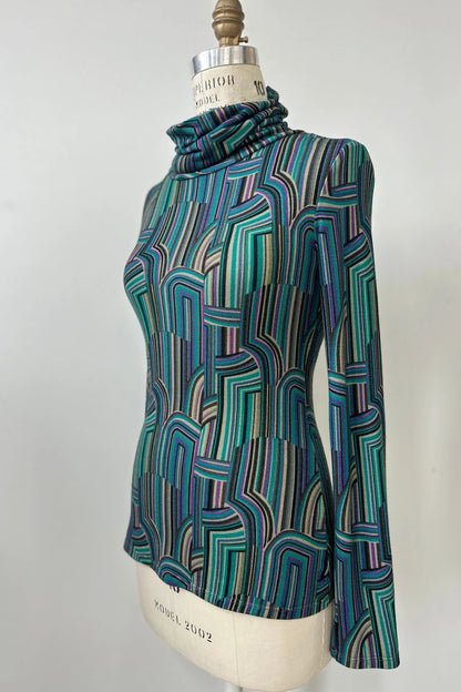 Side view of the Yrene Top by Kollontai in Turquoise, a geometric print turtleneck, is shown on a mannequin against a grey background. 