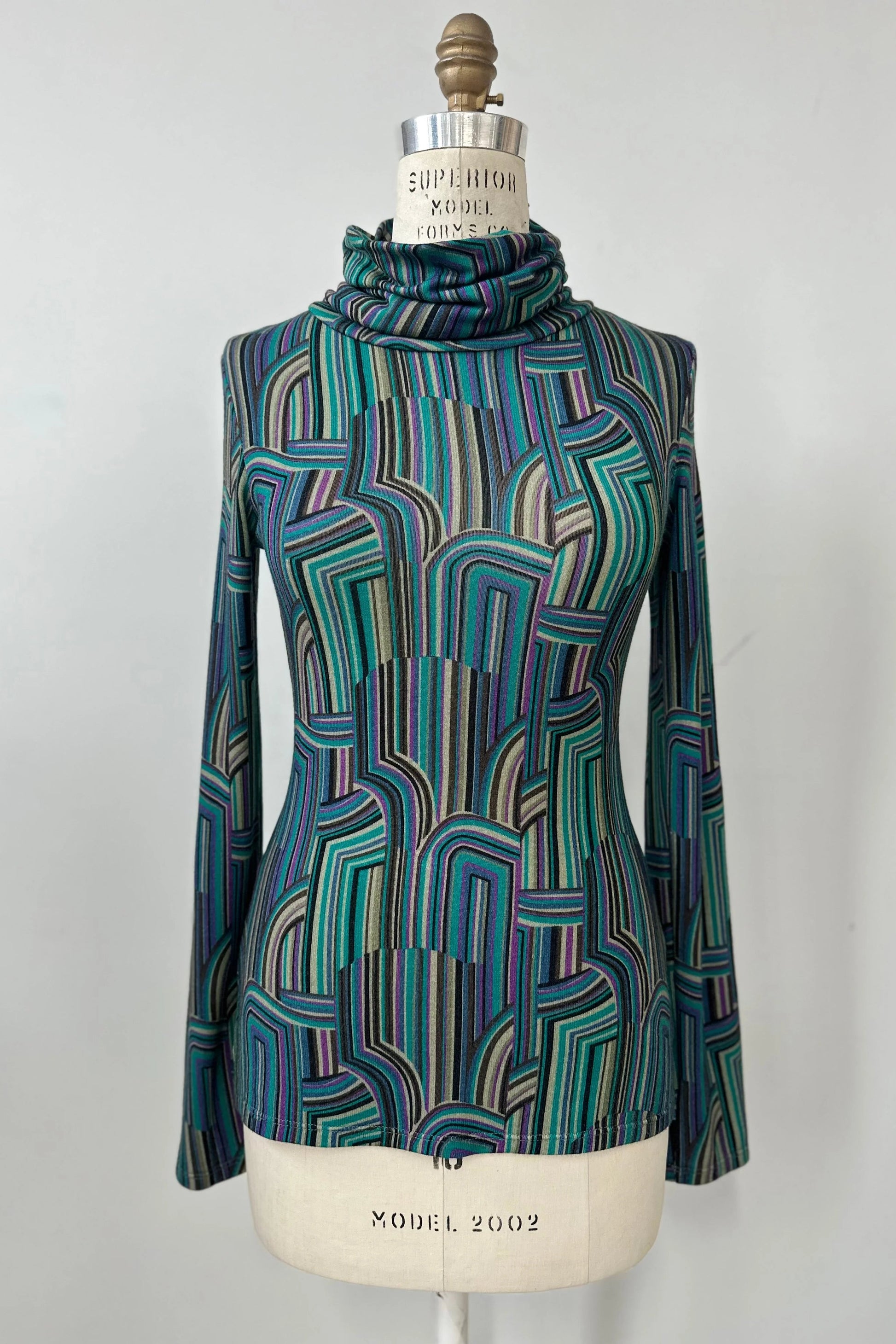 The Yrene Top by Kollontai in Turquoise, a geometric print turtleneck, is shown on a mannequin against a grey background. 