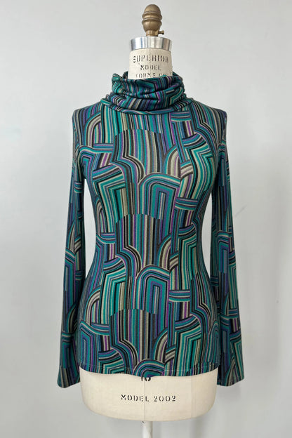 The Yrene Top by Kollontai in Turquoise, a geometric print turtleneck, is shown on a mannequin against a grey background. 