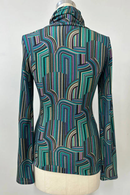 Back view of the Yrene Top by Kollontai in Turquoise, a geometric print turtleneck, is shown on a mannequin against a grey background. 