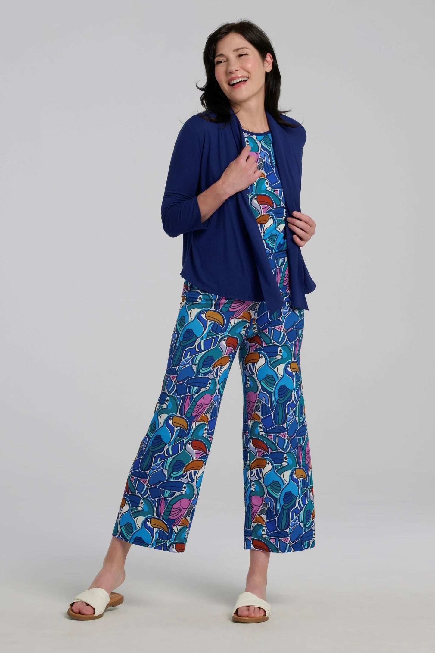 Woman wearing the Hanni Top by Kollontai in Admiral, a cap sleeved relaxed top with a jewel-toned parrot pattern on the front a solid blue back. She is wearing it with a blue bolero and matching pants and standing in front of a grey background. 