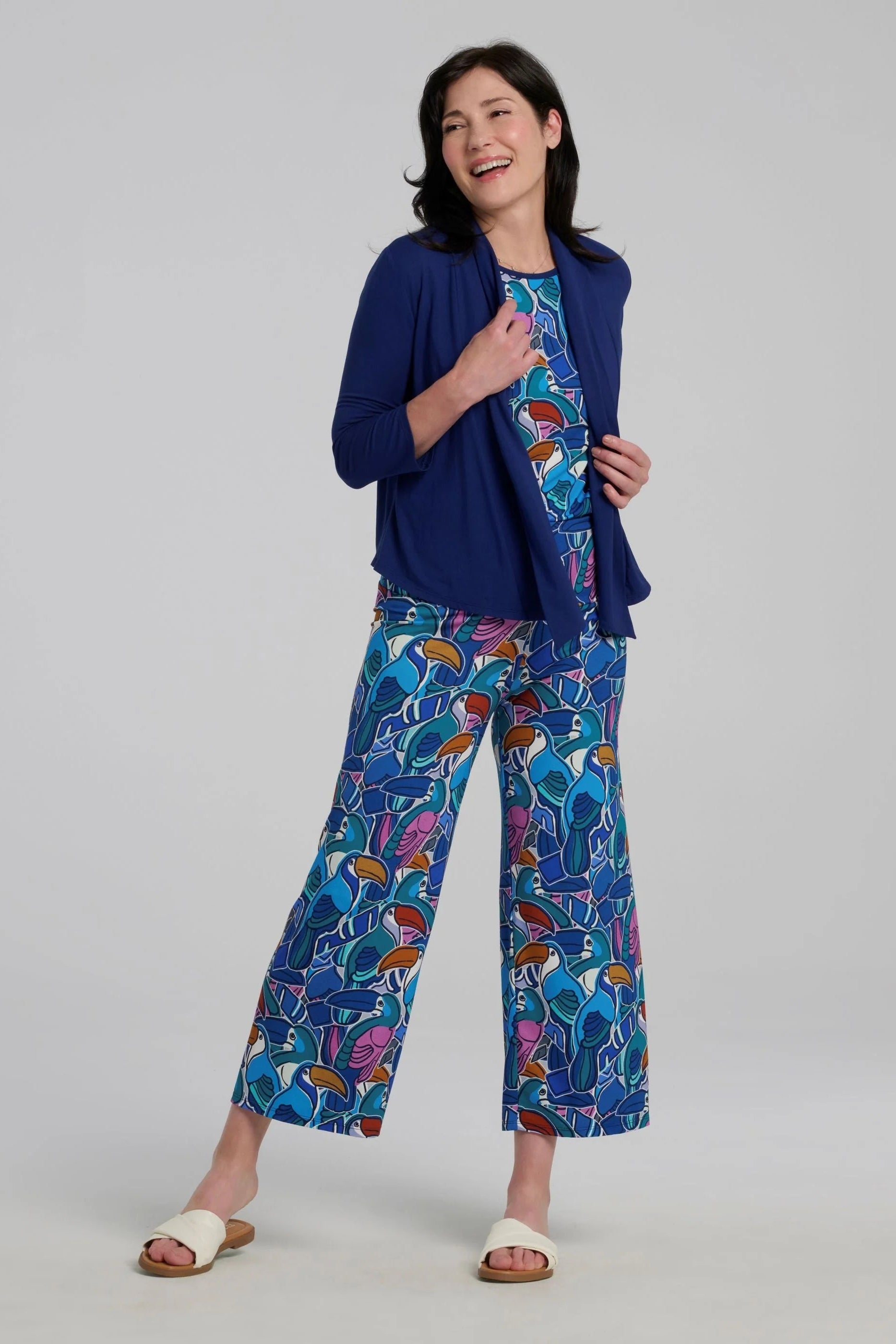 Woman wearing the Hanni Top by Kollontai in Admiral, a cap sleeved relaxed top with a jewel-toned parrot pattern on the front a solid blue back. She is wearing it with a blue bolero and matching pants and standing in front of a grey background. 