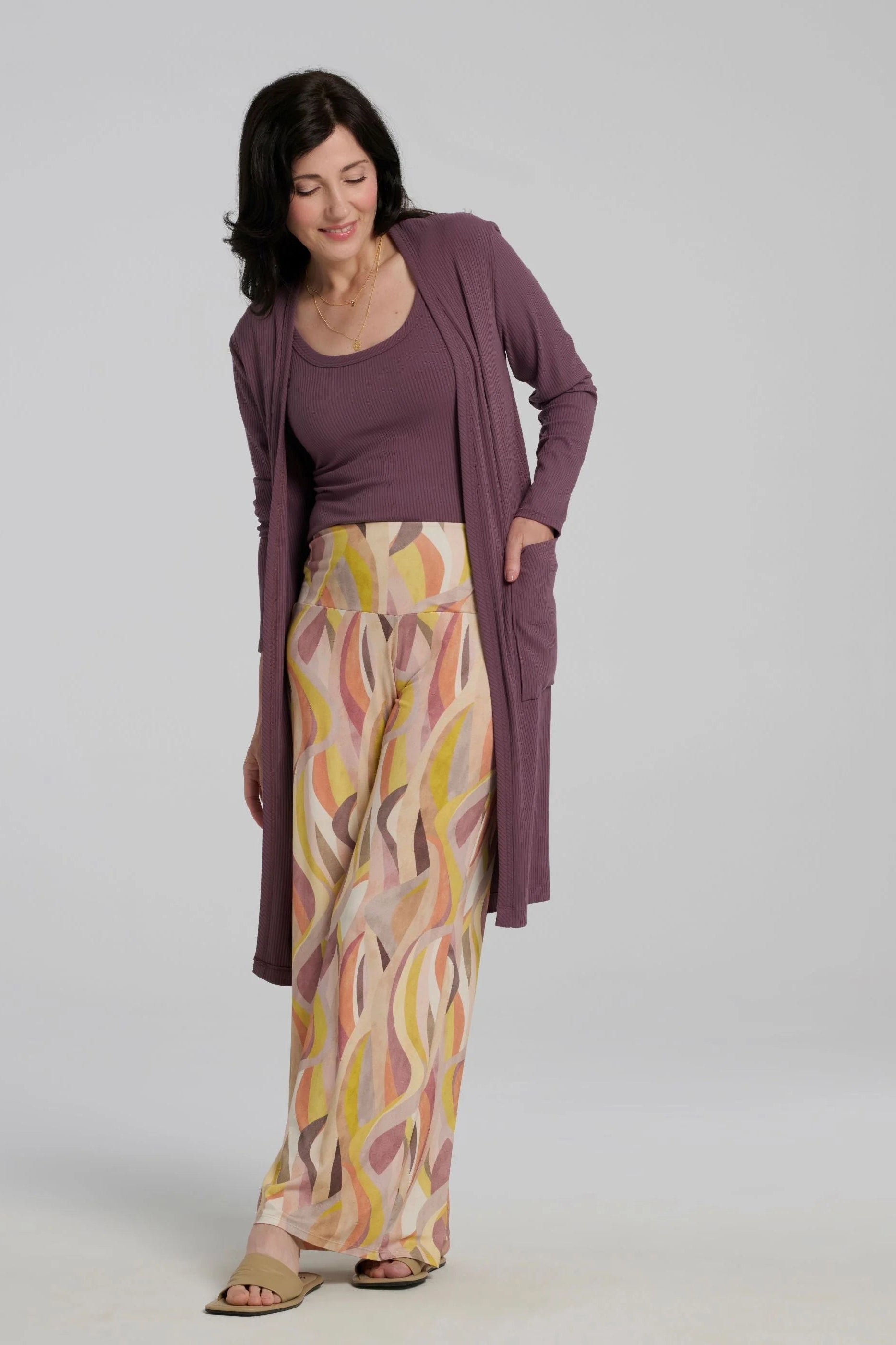 A woman wearing the Sorela pants from Kollontain in Peach print, with a wide pull-on waistband, flowing legs, and swirling geometric print. She is wearing them with a purple tank and duster and standing in front of a white background. 