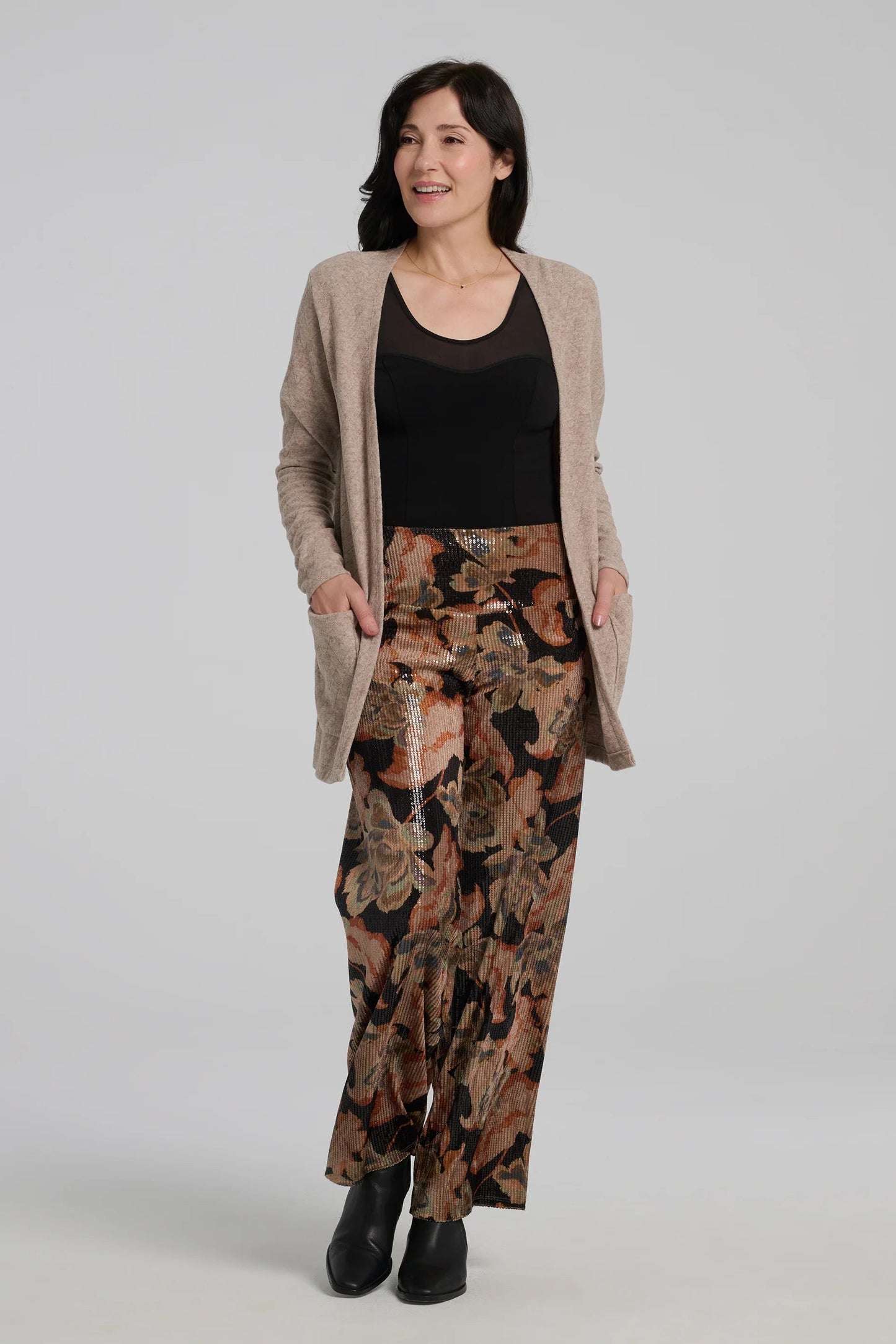 A woman wearing the Carina Pants by Kollontai, featuring floral velvet in autumn tones with clear sequins sewn right into the fabric. They have full-length straight legs and a wide pull-on waistband. She's wearing them with a black tank and beige cardigan and standing in front of a grey background. 