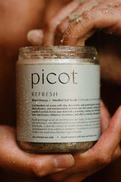 A close-up of a person holding a jar of Picot Refresh Blood Orange + Menthol Salt Scrub, with sand visible on the jar and fingers, against a warm, blurred skin-toned background.