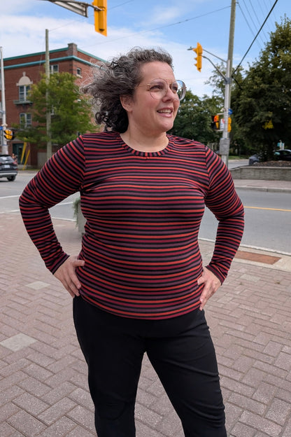 A woman wearing the A25 Sweater by Slak in Colourful Stripe, a long sleeved top with a round neck and a slim fit. She is wearing it with black pants and standing on the street. 