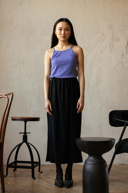 A woman wearing the Simone Skirt by Lights of All in Black, a sleek calf-length skirt with a flared hem and an elastic waist. She is wearing it with a purple top and standing in a room with furniture. 