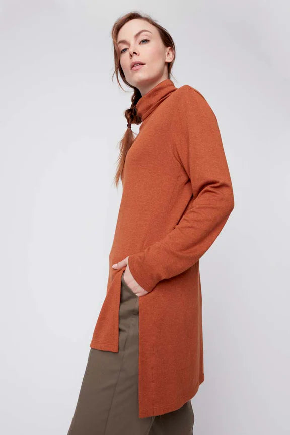 A woman wearing the Melanie Tunic by Julei in Ochre, a long sleeved turtleneck tunic with a hi-low hemline and side slits. She is wearing it with green pants and standing in front of a white background. 