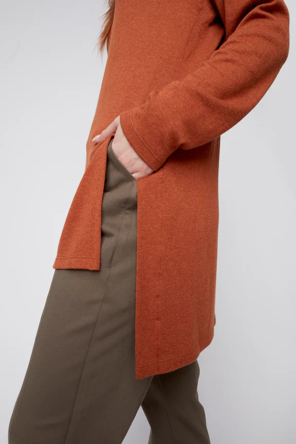 Close up side view of a woman wearing the Melanie Tunic by Julei in Ochre, a long sleeved turtleneck tunic with a hi-low hemline and side slits. She is wearing it with green pants and standing in front of a white background. 