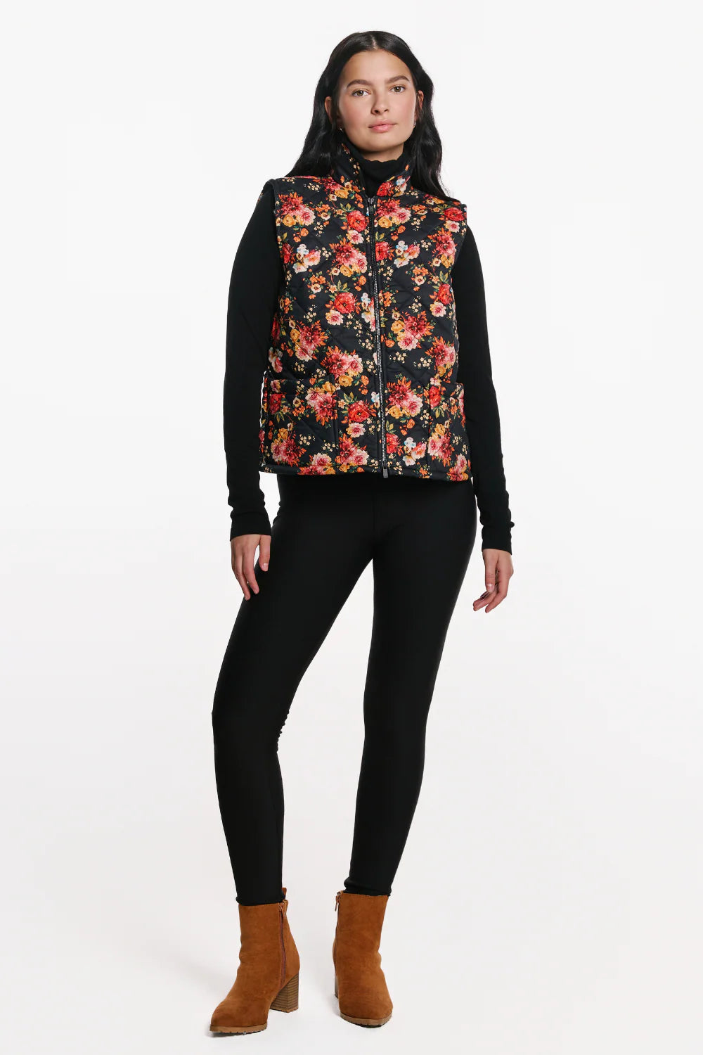 A woman wearing the Harper Vest by EMK, a quilted floral vest with a zip front two pockets, and black waffle knit fabric at the collar. She is wearing it with black pants and standing in front of a white background. 