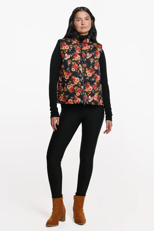 A woman wearing the Harper Vest by EMK, a quilted floral vest with a zip front two pockets, and black waffle knit fabric at the collar. She is wearing it with black pants and standing in front of a white background. 