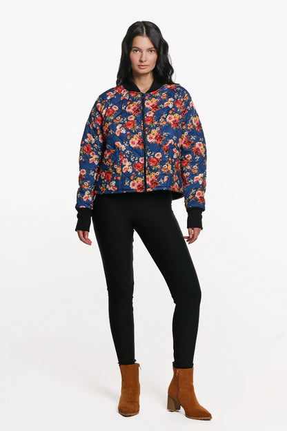 A woman wearing the Harper Bomber Jacket by EMK, a floral bomber jacket in quilted fabric with black waffle knit fabric at the cuffs and collar. She is wearing it with black pants and standing in front of a white background. 