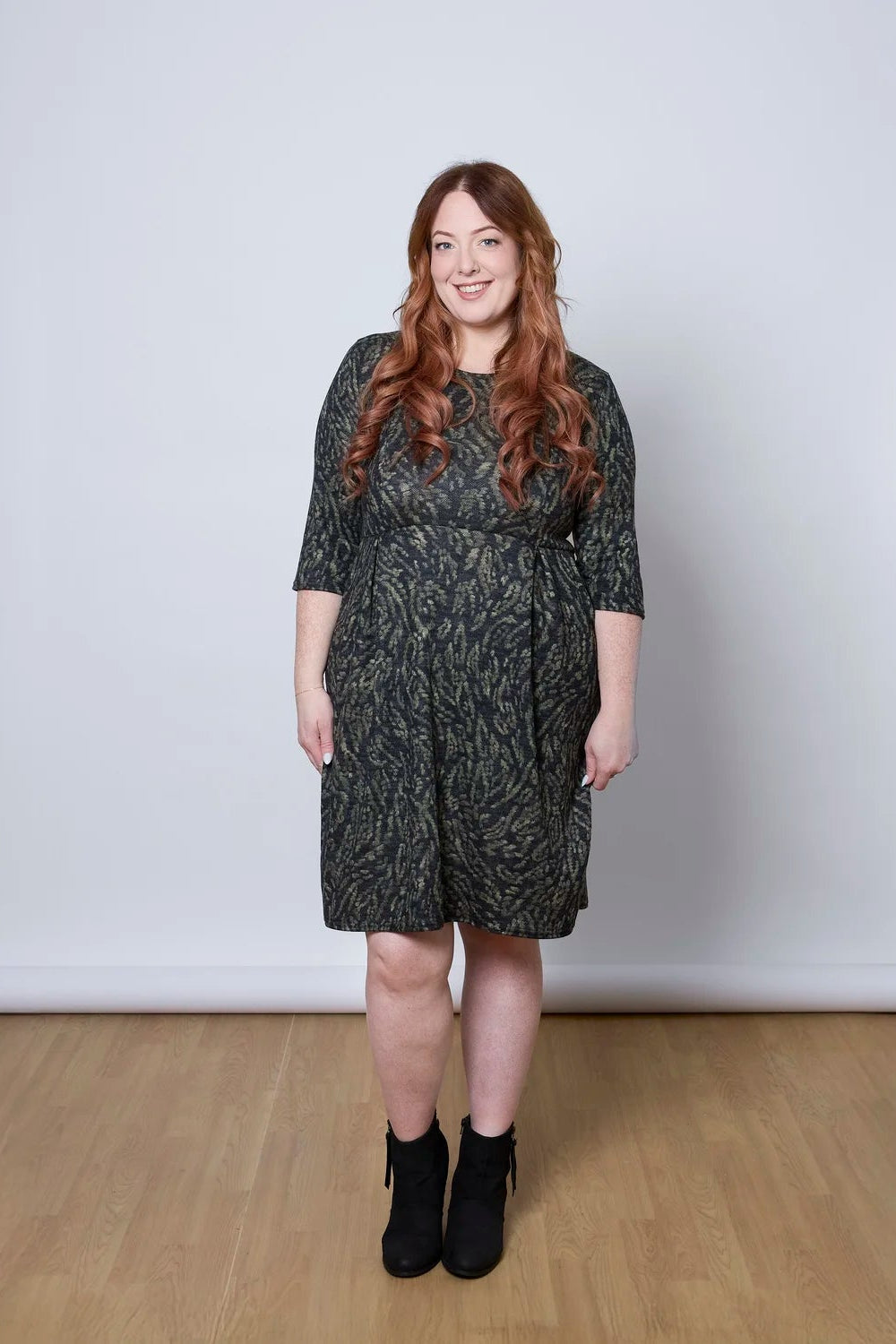A woman wearing the Meadow Dress by Tangente in Green Print, with a round neck, 3/4 sleeves, princess seams, and a pleated knee-length skirt. She is standing against a white wall. 