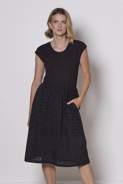 A woman wearing the Primrose Dress by Tangente in Black, a knee-length dress featuring a bamboo rayon bodice and daisy eyelet skirt, with short sleeves and a round neck, standing against a plain background. 