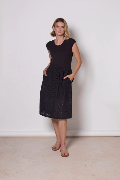 A woman wearing the Primrose Dress by Tangente in Black, a knee-length dress featuring a bamboo rayon bodice and daisy eyelet skirt, with short sleeves and a round neck, standing against a plain background. 