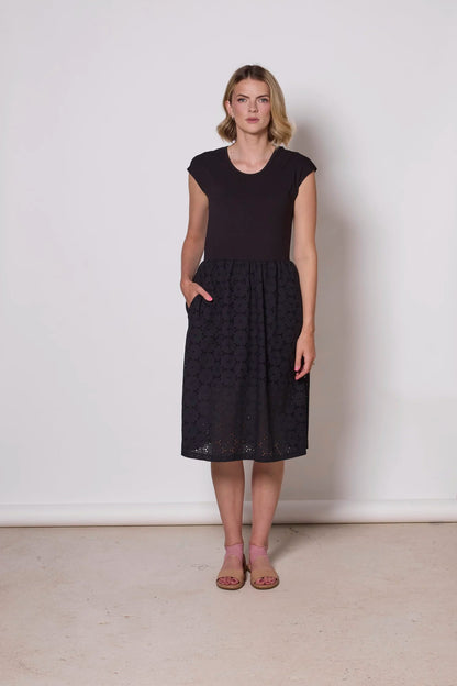 A woman wearing the Primrose Dress by Tangente in Black, a knee-length dress featuring a bamboo rayon bodice and daisy eyelet skirt, with short sleeves and a round neck, standing against a plain background. 