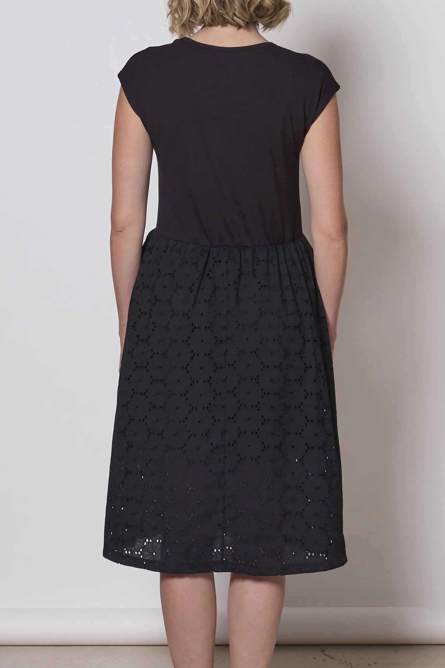 Back view of a woman wearing the Primrose Dress by Tangente in Black, a knee-length dress featuring a bamboo rayon bodice and daisy eyelet skirt, with short sleeves and a round neck, standing against a plain background. 