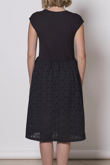 Back view of a woman wearing the Primrose Dress by Tangente in Black, a knee-length dress featuring a bamboo rayon bodice and daisy eyelet skirt, with short sleeves and a round neck, standing against a plain background. 