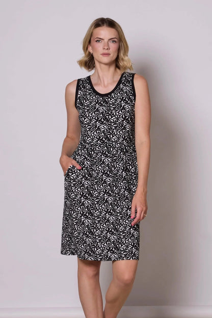 A woman wearing the Mai Dress by Tangente in Black and White Floral print, a sleeveless bamboo jersey dress with a round neck and  gathered knee-length skirt. She is standing in front of a grey background.  