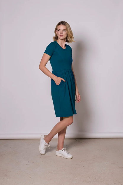 A woman wearing the Hayley Dress by Tangente in Teal, a short sleeved bamboo dress with a gathered knee-length skirt. She is standing in front of a grey background.