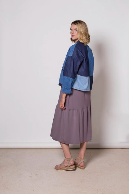 A woman wearing the Myrtle Denim Jacket by Tangente, a patchwork jacket with an open front, drop sleeves, and front patch pockets. She is wearing it over a purple dress against a white background