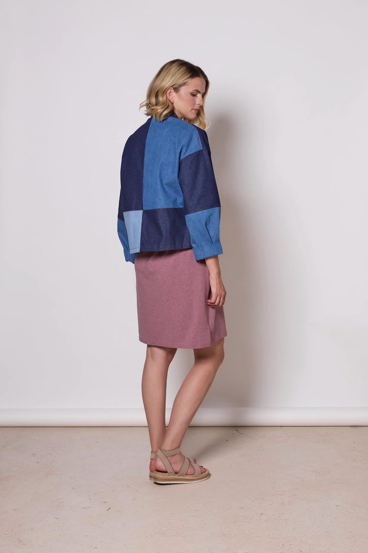 Back view of a woman wearing the Myrtle Denim Jacket by Tangente, a patchwork jacket with an open front, drop sleeves, and front patch pockets. She is wearing it over a pink dress against a white background