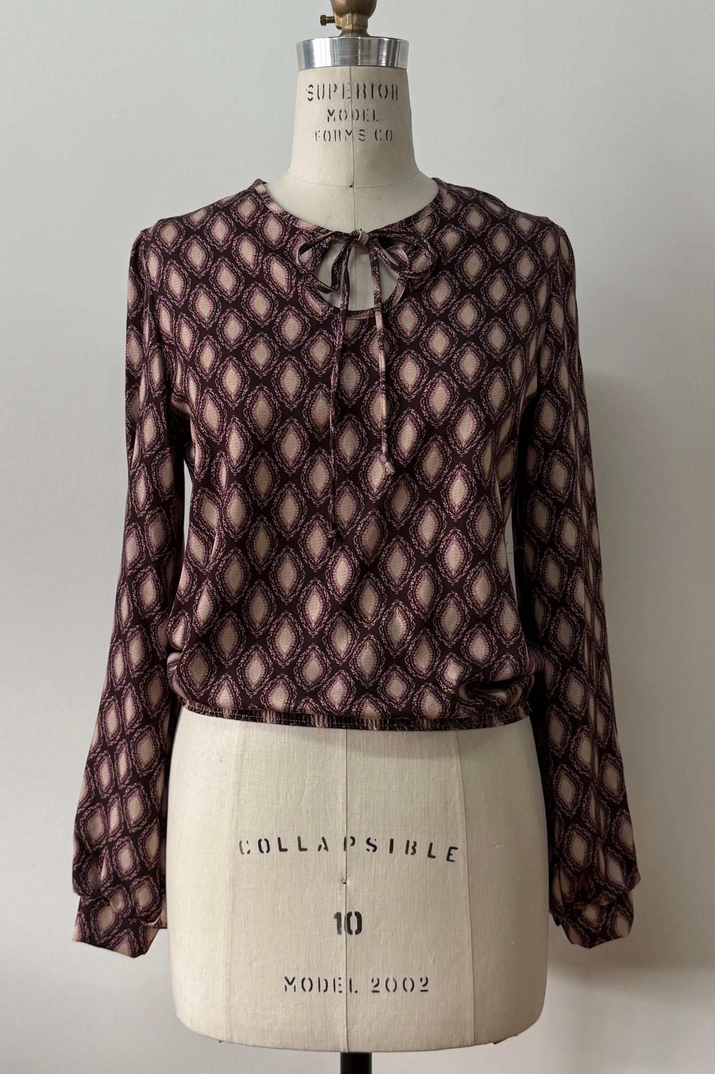  The Isabella Blouse by Kollontai, a burgundy and pink patterned blouse with a tie detail at the neck, balloon sleeves with square jewel buttons, and an elastic waist, is shown on a mannequin against a white background. 