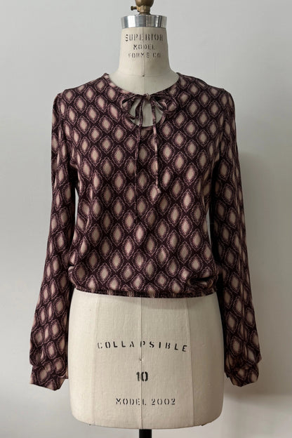  The Isabella Blouse by Kollontai, a burgundy and pink patterned blouse with a tie detail at the neck, balloon sleeves with square jewel buttons, and an elastic waist, is shown on a mannequin against a white background. 