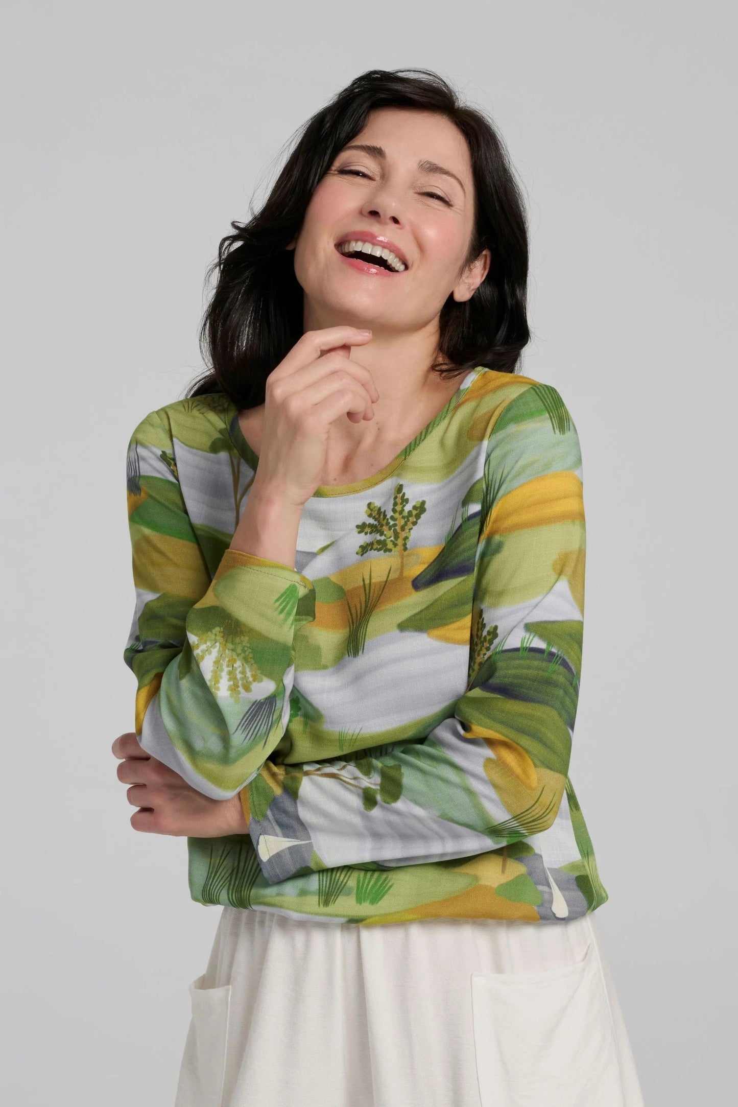 A woman wearing the Belica Blouse by Kollontai in a Granny Smith toned nature print. It has a wide neck, long sleeves, linen-look fabric, and an elasticized waist for a subtle blouson effect She is wearing it with white pants and standing in front of a grey background. 