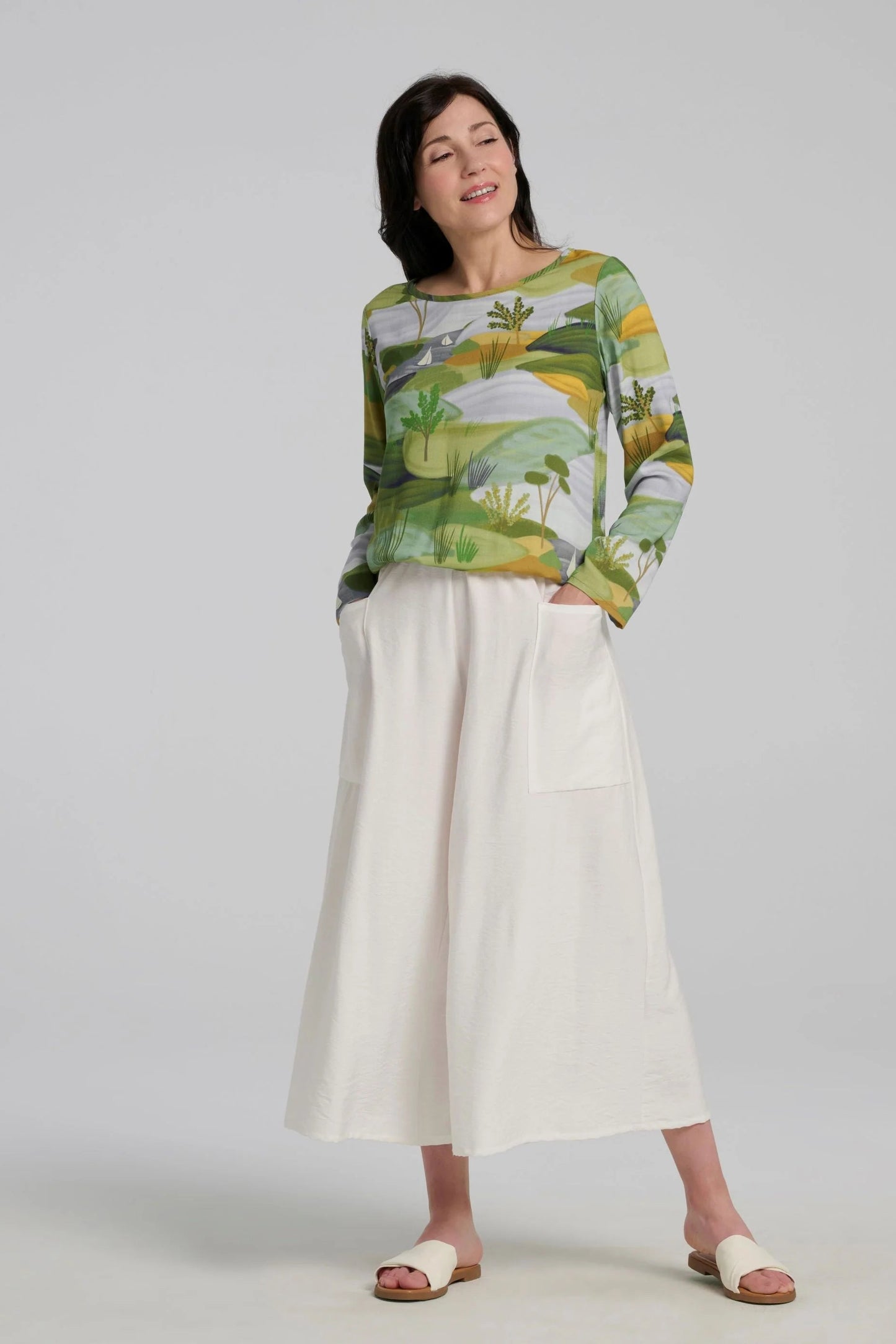 A woman wearing the Belica Blouse by Kollontai in a Granny Smith toned nature print. It has a wide neck, long sleeves, linen-look fabric, and an elasticized waist for a subtle blouson effect She is wearing it with the Ishtar pants in white pants and standing in front of a grey background. 