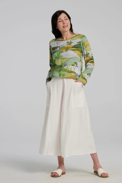 A woman wearing the Belica Blouse by Kollontai in a Granny Smith toned nature print. It has a wide neck, long sleeves, linen-look fabric, and an elasticized waist for a subtle blouson effect She is wearing it with the Ishtar pants in white pants and standing in front of a grey background. 