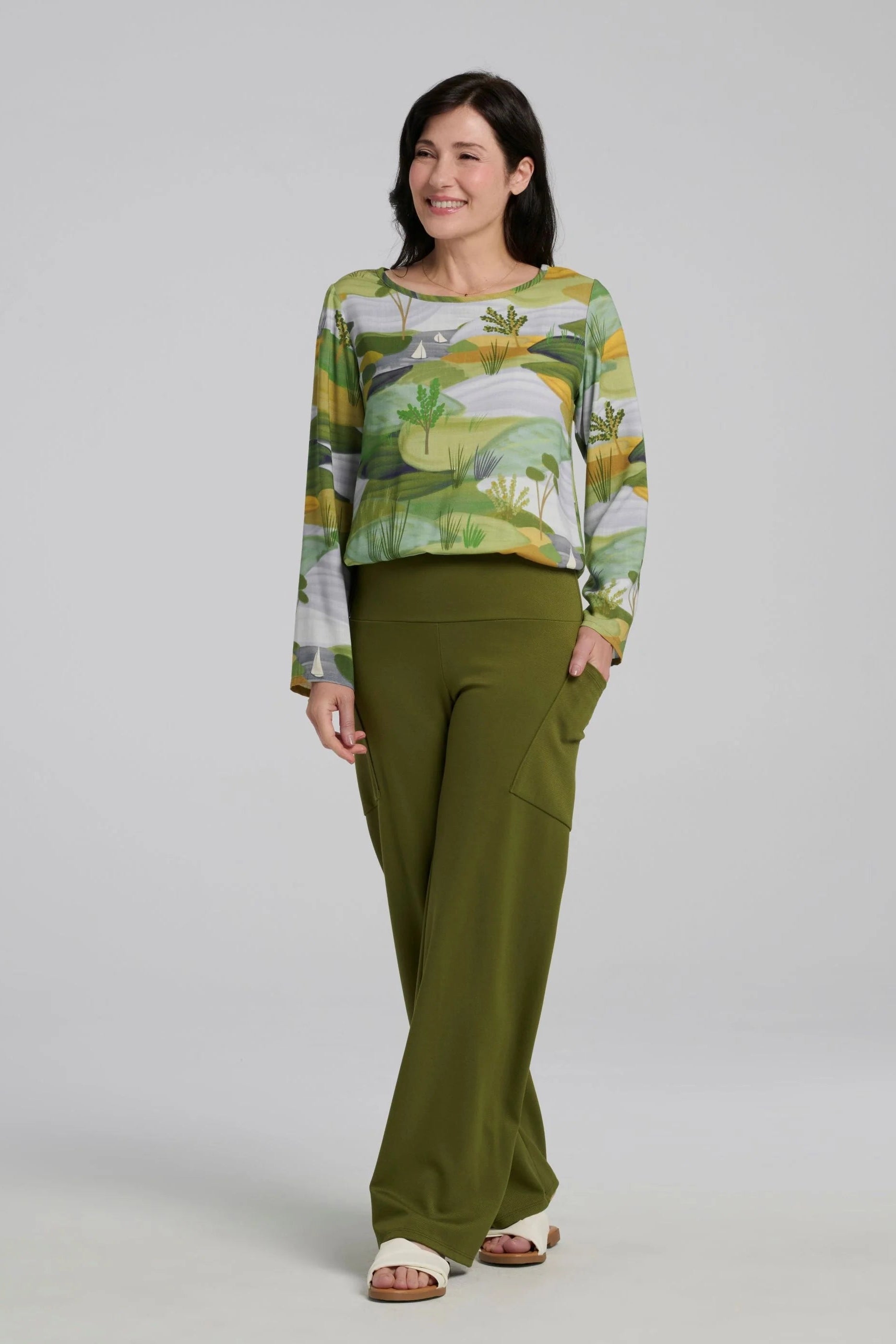 A woman wearing the Belica Blouse by Kollontai in a Granny Smith toned nature print. It has a wide neck, long sleeves, linen-look fabric, and an elasticized waist for a subtle blouson effect She is wearing it with the Susan Pants in Granny Smith and standing in front of a grey background. 