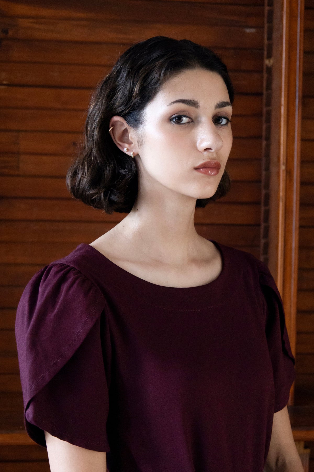 Close up view of a woman wearing the Opal Dress by Eve Lavoie in Burgundy, a short dress with tulip sleeves, and an A-line shape. She's standing indoors by a wood panelled wall. 