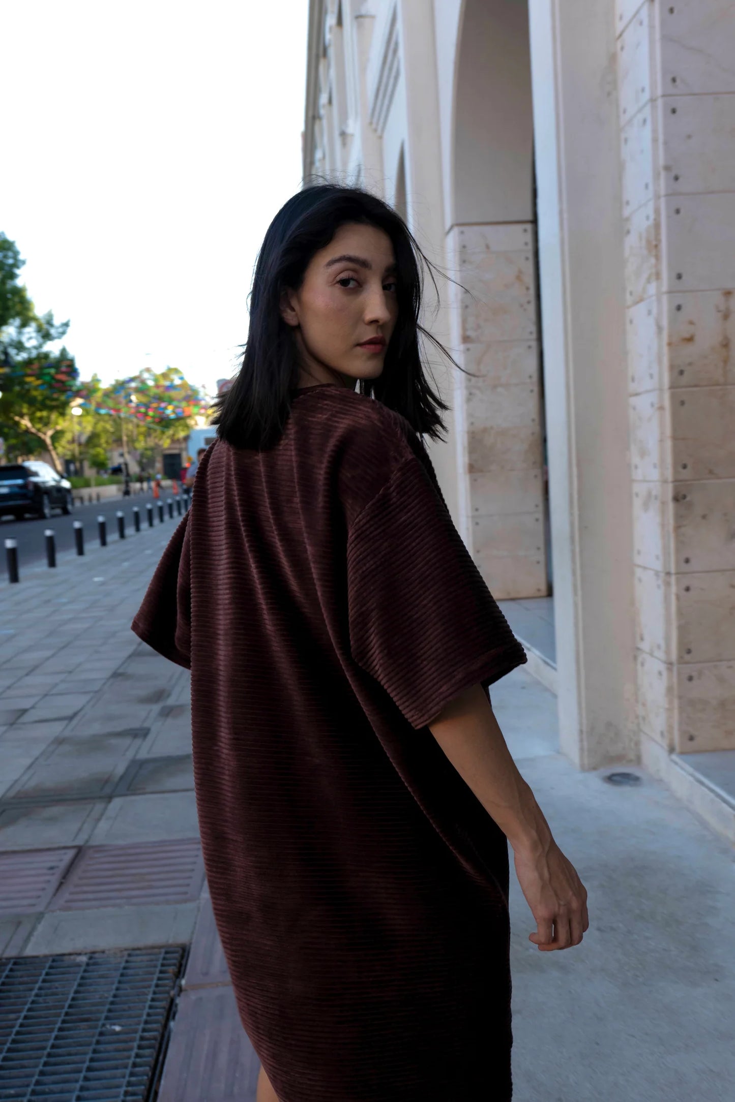 A woman wearing the Mykonos Dress by MAS in Cacao, a short sleeved boxy, knee-length dress in ribbed velvet fabric. She is walking down a city street. 