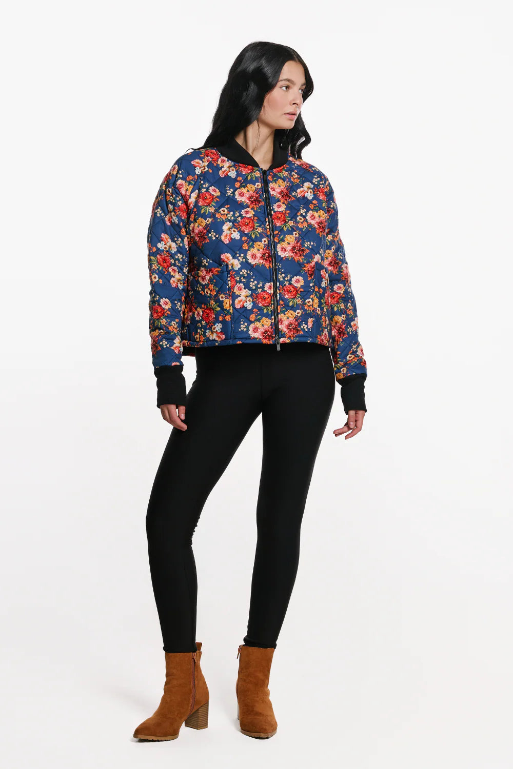 A woman wearing the Harper Bomber Jacket by EMK, a floral bomber jacket in quilted fabric with black waffle knit fabric at the cuffs and collar. She is wearing it with black pants and standing in front of a white background. 