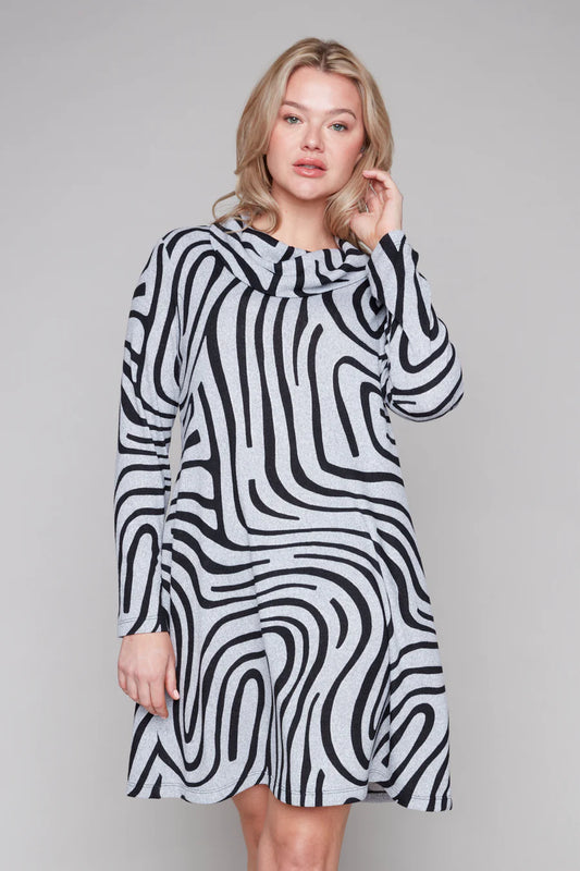 A woman wearing the Powell Dress by Compli K in Black and Silver Swirl, featuring a cowl neck, long sleeves, and a slight trapeze shape. She is standing in front of a grey background. 