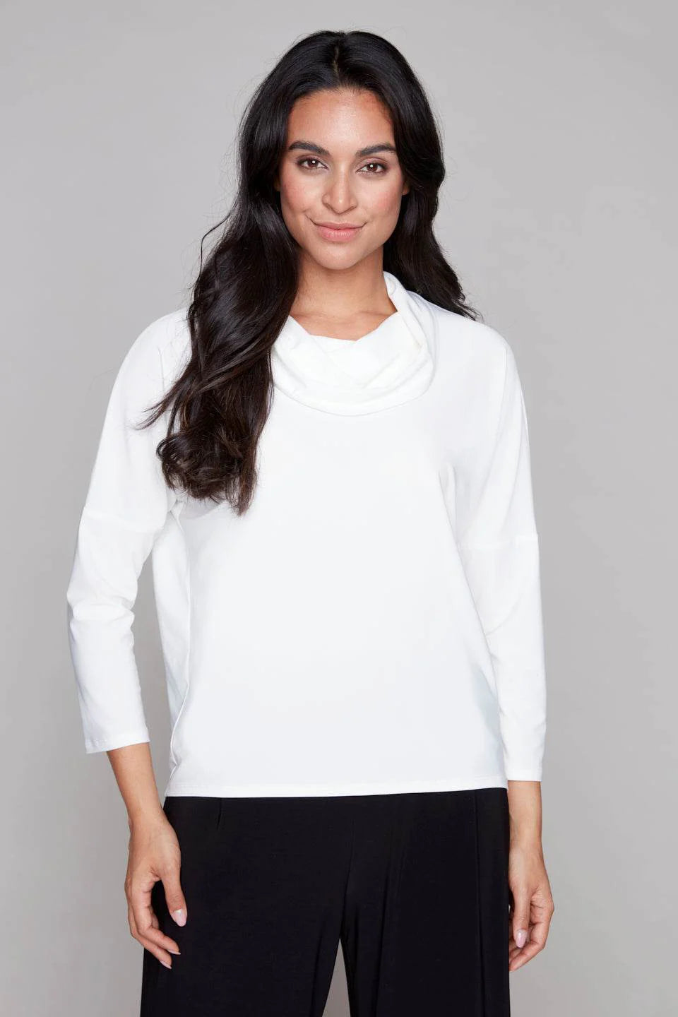 A woman wearing the Perry Top by Compli K in Ivory, featuring a cowl neck, 3/4 drop sleeves, and decorative buttons up the back. She is wearing it with black pants and standing in front of a white background. 