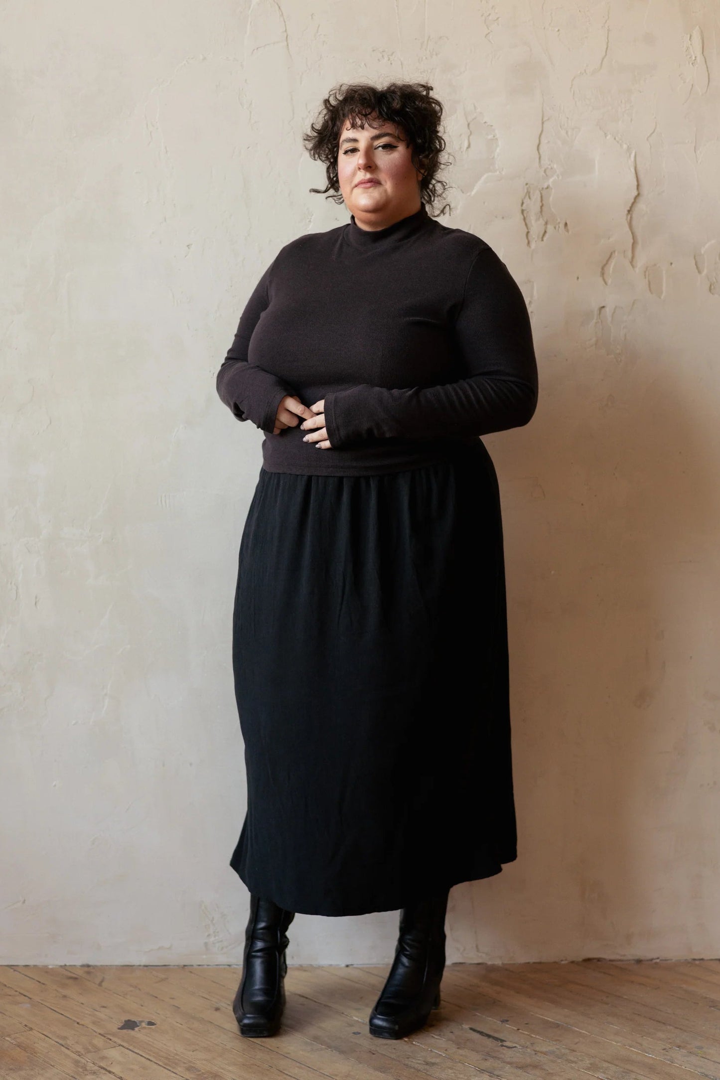A woman wearing the Simone Skirt by Lights of All in Black, a sleek calf-length skirt with a flared hem and an elastic waist. She is wearing it with a brown turtleneck and standing by a beige wall. 