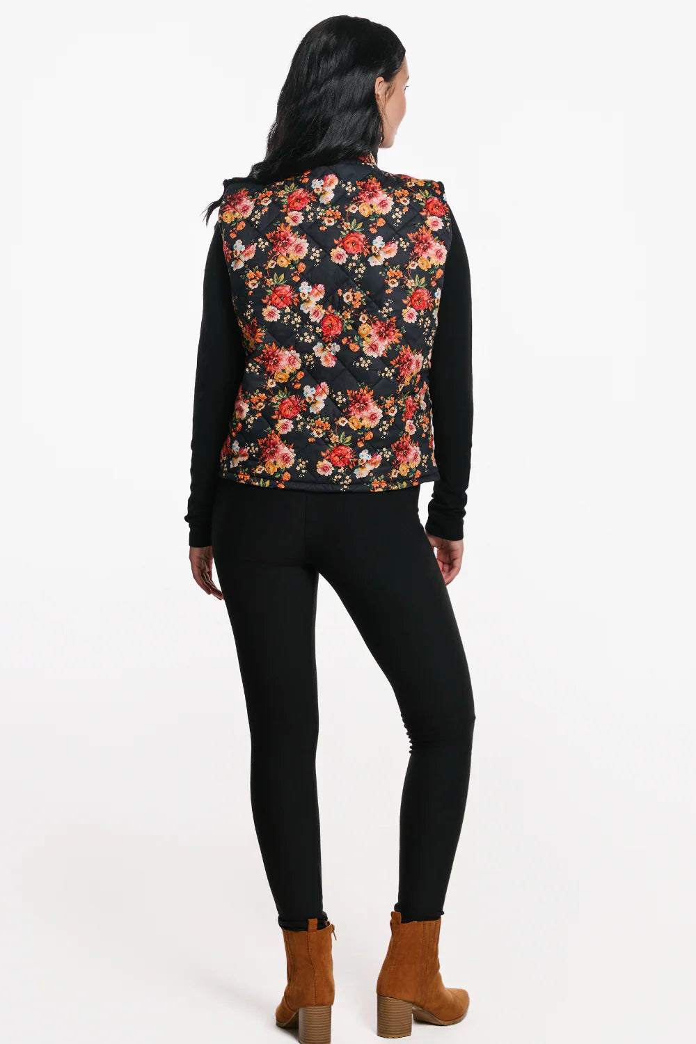 Back view of a woman wearing the Harper Vest by EMK, a quilted floral vest with a zip front two pockets, and black waffle knit fabric at the collar. She is wearing it with black pants and standing in front of a white background. 