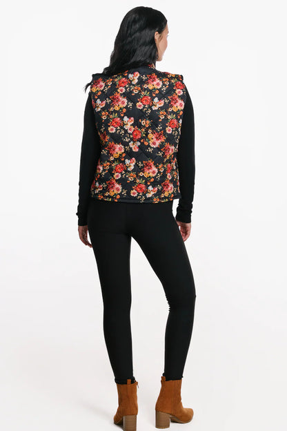 Back view of a woman wearing the Harper Vest by EMK, a quilted floral vest with a zip front two pockets, and black waffle knit fabric at the collar. She is wearing it with black pants and standing in front of a white background. 