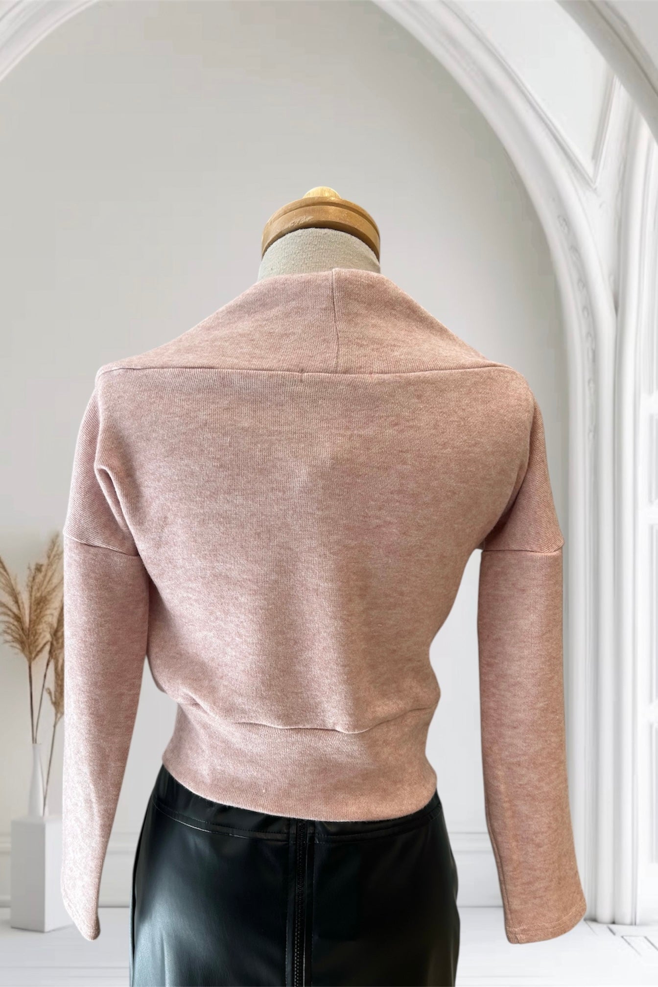 A back view of the Changes Sweater by Allison Wonderland in Pink, featuring a large cowl neck, long dropped sleeves, and a wide waistband, is shown with the Desire Skirt in Black against a white arched backdrop. 