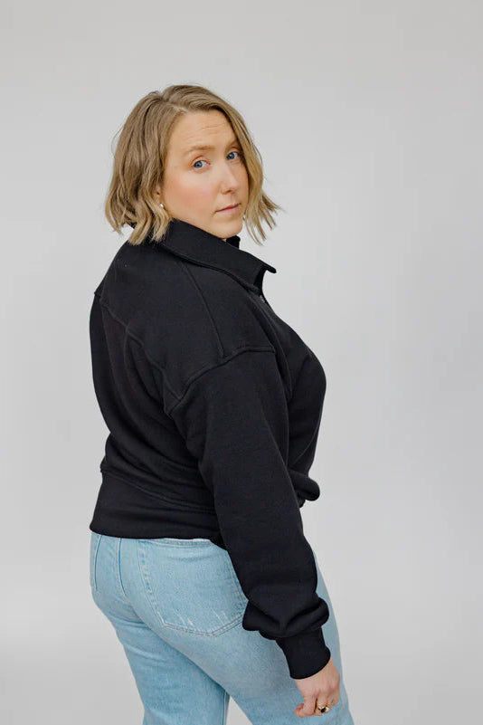 Back view of a woman wearing the Classic Coastal Sweater by Blondie in Black, a relaxed fit sweatshirt with a collar and four button placket. She is wearing it with jeans and standing in front of a white background. 