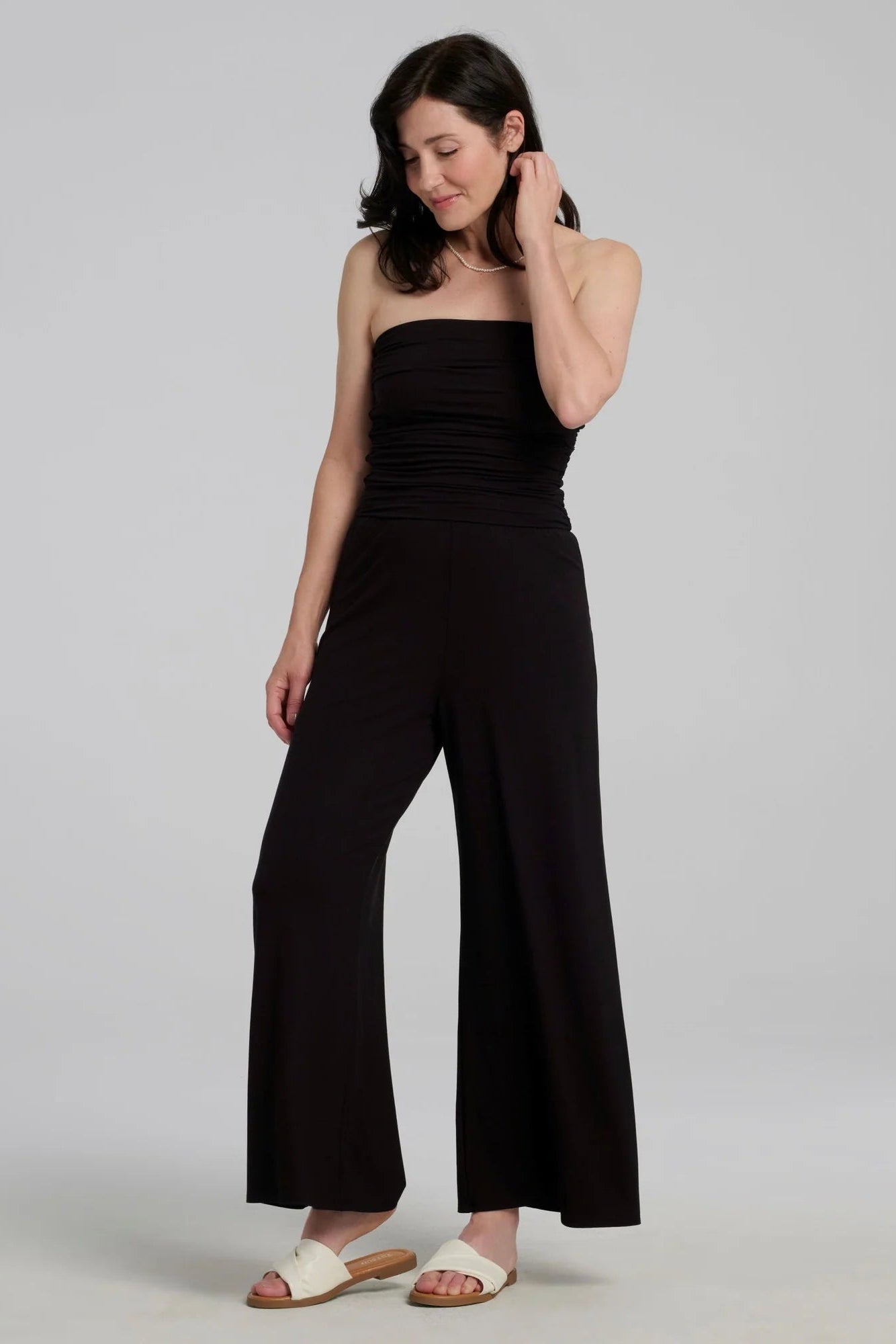 A woman wearing the black strapless Dolora Jumpsuit from Kollontai, with wide palazzo legs, standing against a grey background.