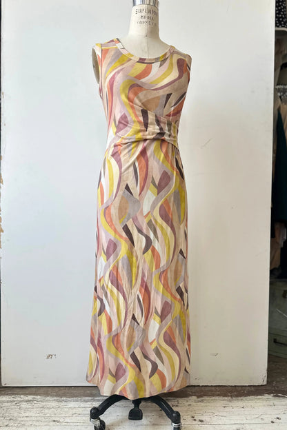 The Serina Dress by Kollontai in Peach print, a long, slim cut sleeveless dress with a soft pleat at the waist, is shown on a mannequin in front of a white background. 