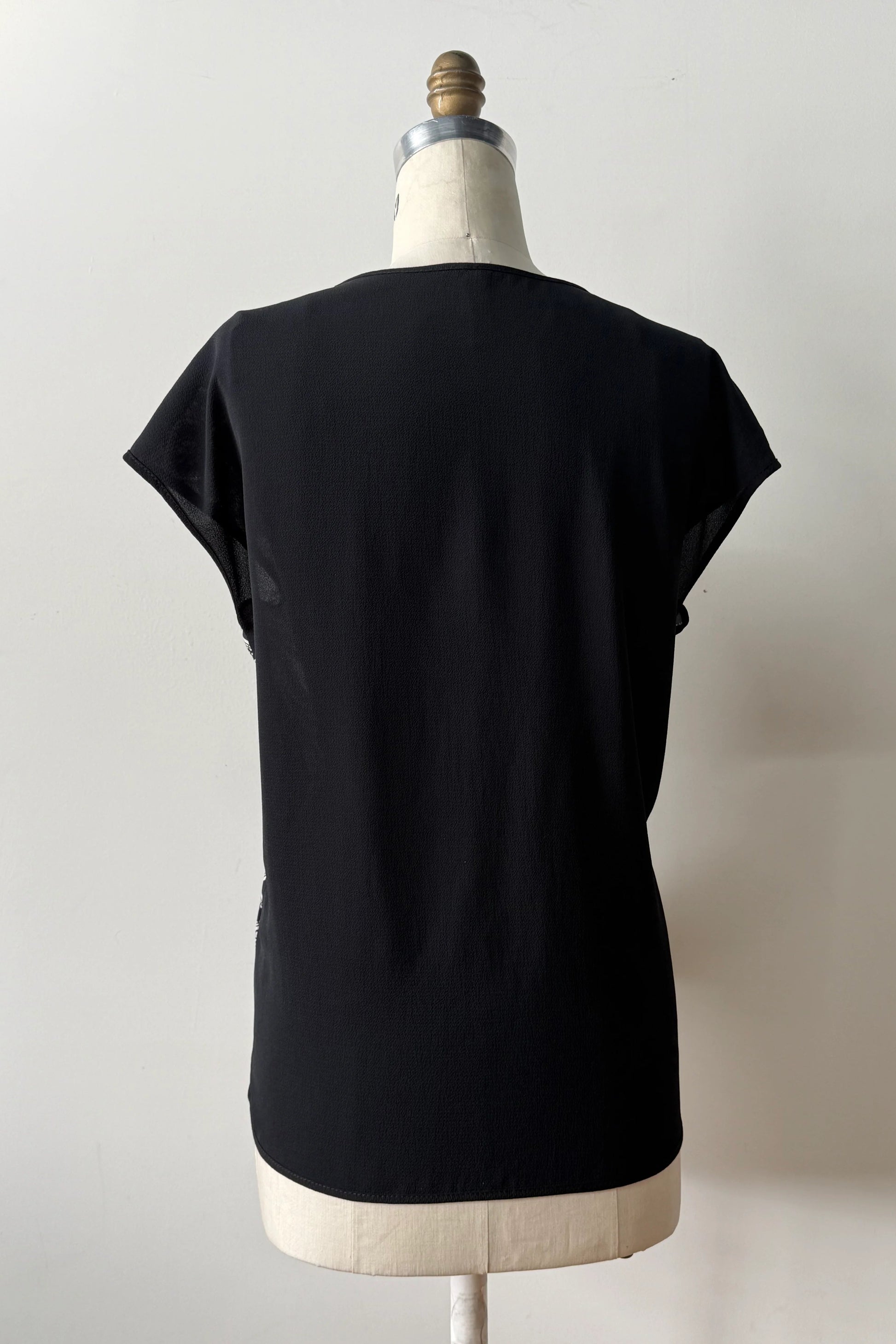 Back view of the Malo T-shirt from Kollontai, featuring a floral and panther print in black-on-ivory on the front, cap sleeves, and a solid black back, shown on a mannequin in front of a grey background 