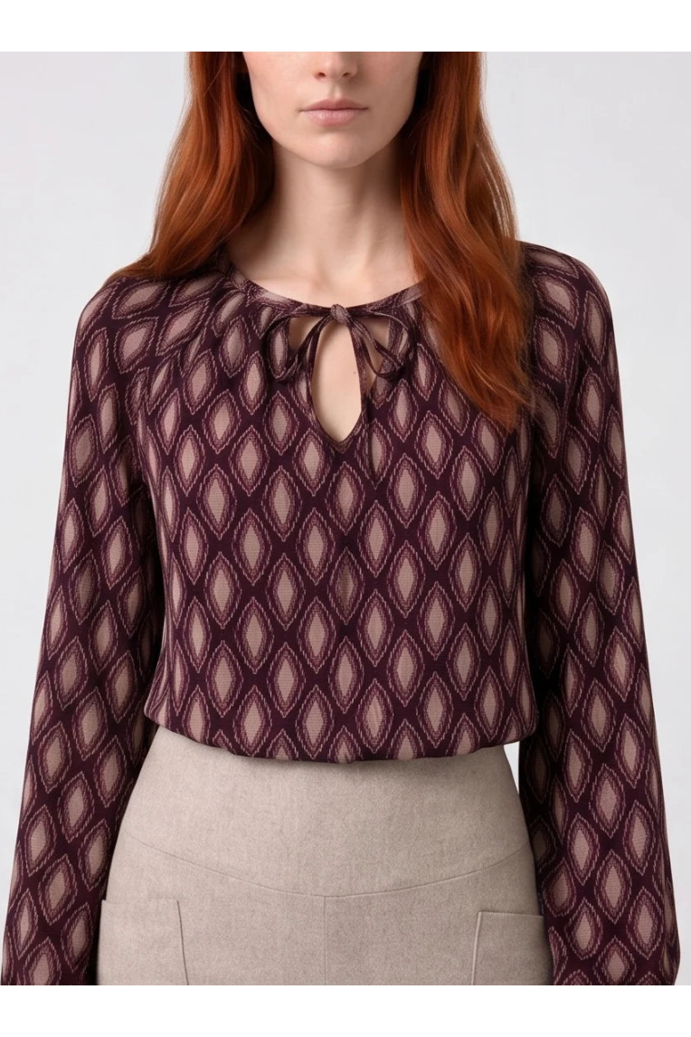 Torso view of a woman wearing the Isabella Blouse by Kollontai, a burgundy and pink patterned blouse with a tie detail at the neck, balloon sleeves with square jewel buttons, and an elastic waist. She is wearing it with beige pants and standing in front of a white background. 