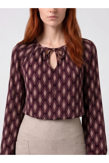 Torso view of a woman wearing the Isabella Blouse by Kollontai, a burgundy and pink patterned blouse with a tie detail at the neck, balloon sleeves with square jewel buttons, and an elastic waist. She is wearing it with beige pants and standing in front of a white background. 