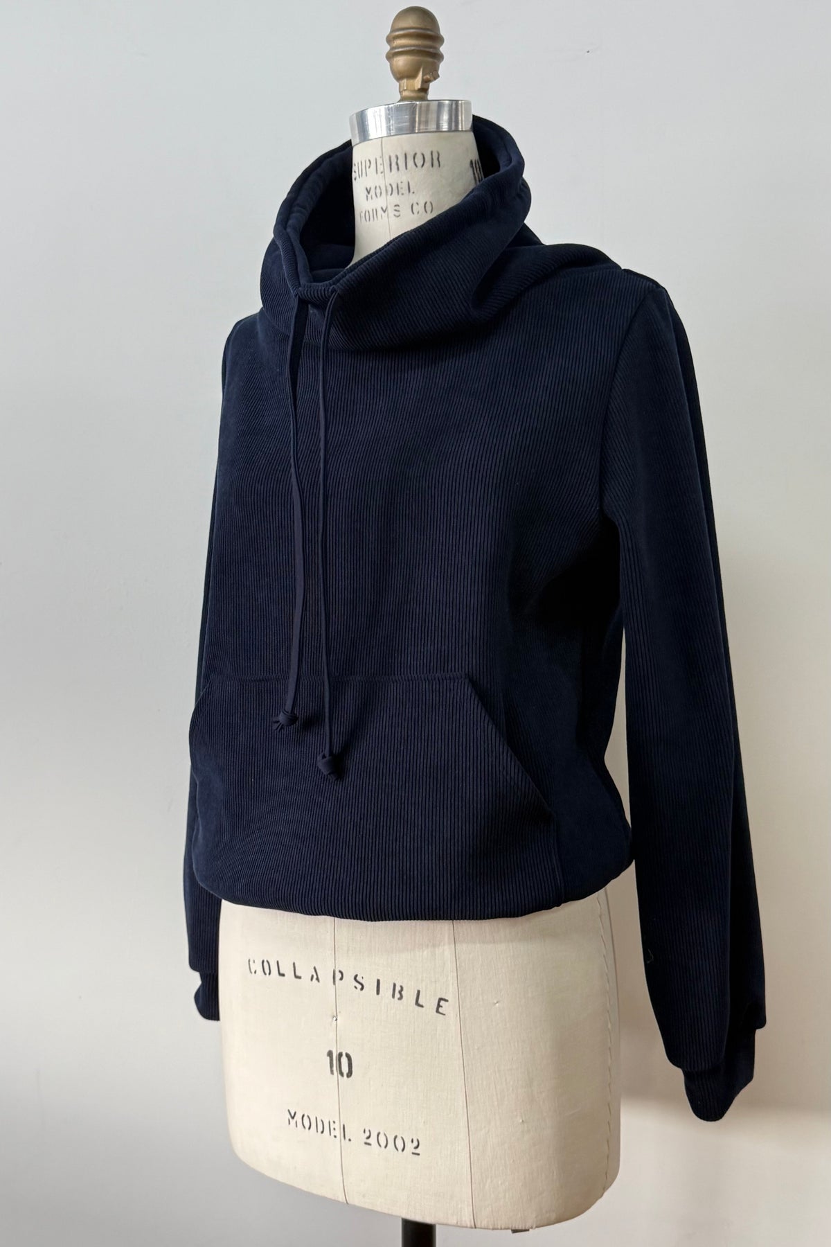 The Ramos Sweater by Kollonta in a ribbed navy corduroy-like fabric, with a large cowl neck with a drawstring and a kangaroo pocket, is shown on a mannequin against a white background. 